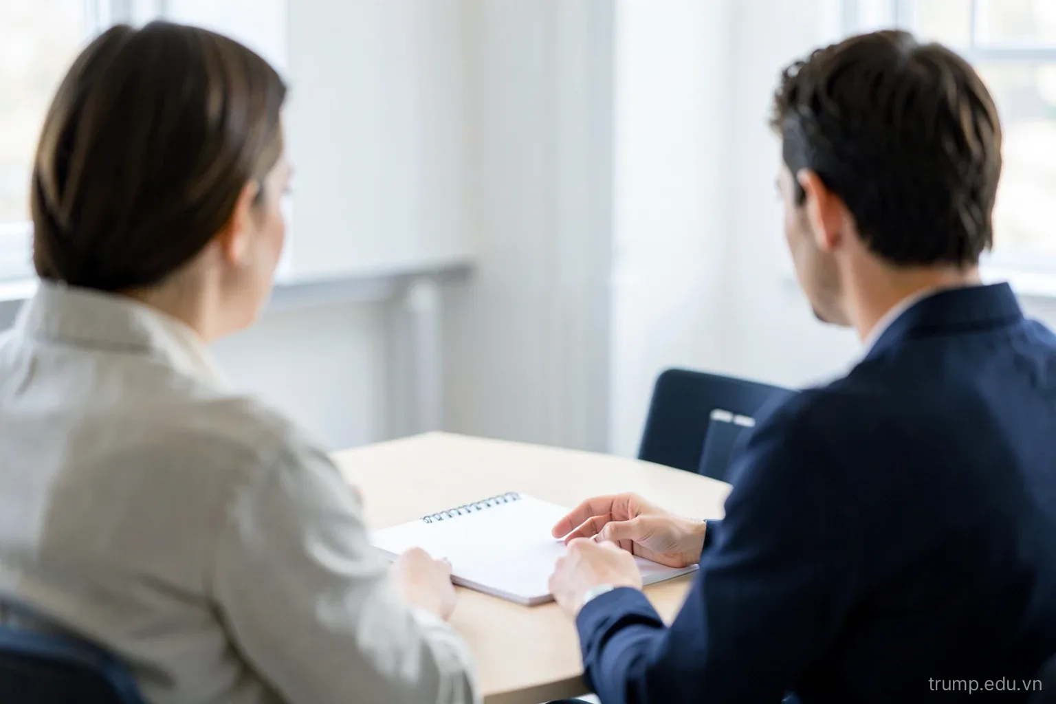 Two people talking across a small meeting table in a bright office, one gesturing toward a blank notepad, over-the-shoulder view, faces not visible, soft white and navy tones