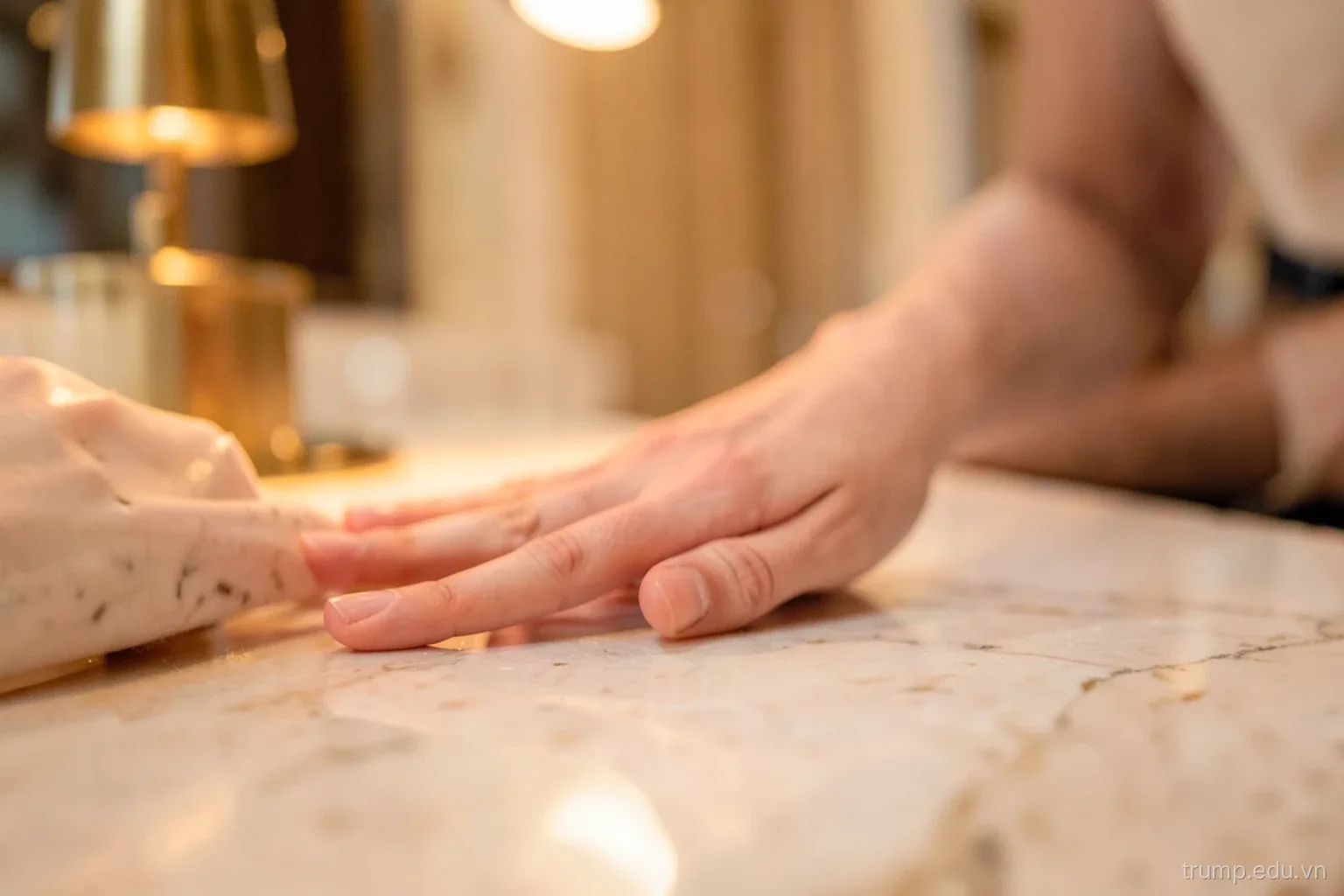 Close-up of a hand touching a polished marble surface in a hotel lobby, warm light from a brass lamp reflecting, shallow depth of field, rose and cream tones